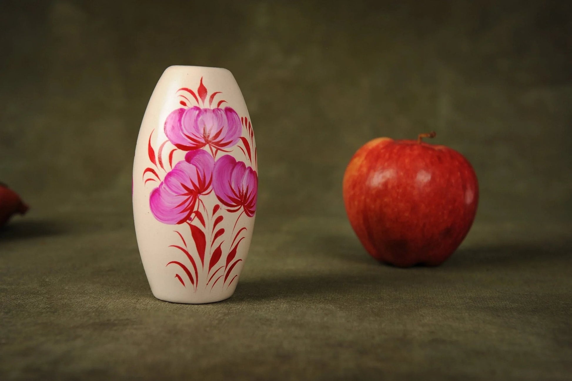 Hand-painted ceramic vase with pink and red floral design beside a red apple on green cloth