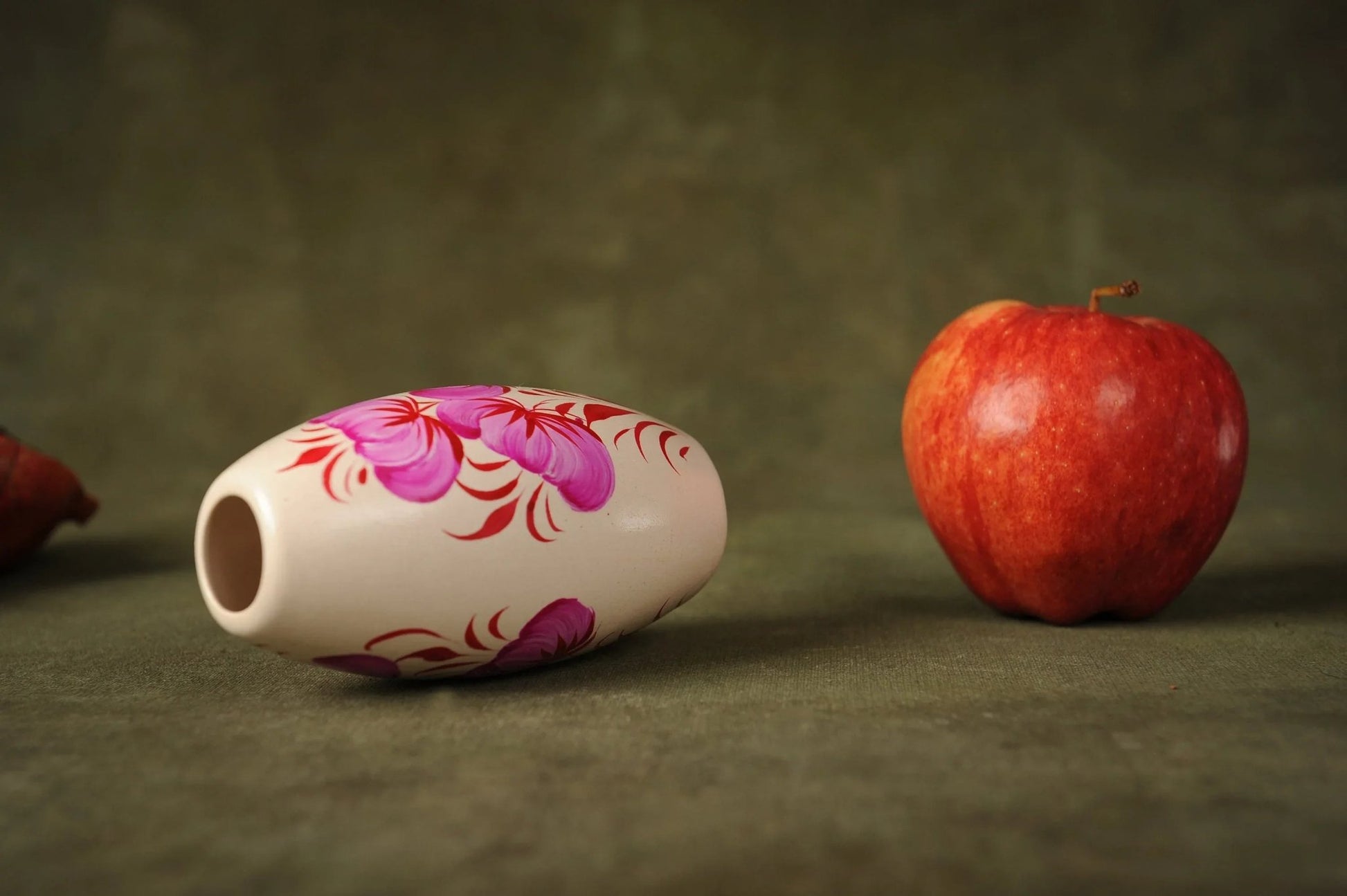 Hand-painted small ceramic vase with pink floral design beside a red apple on a green surface