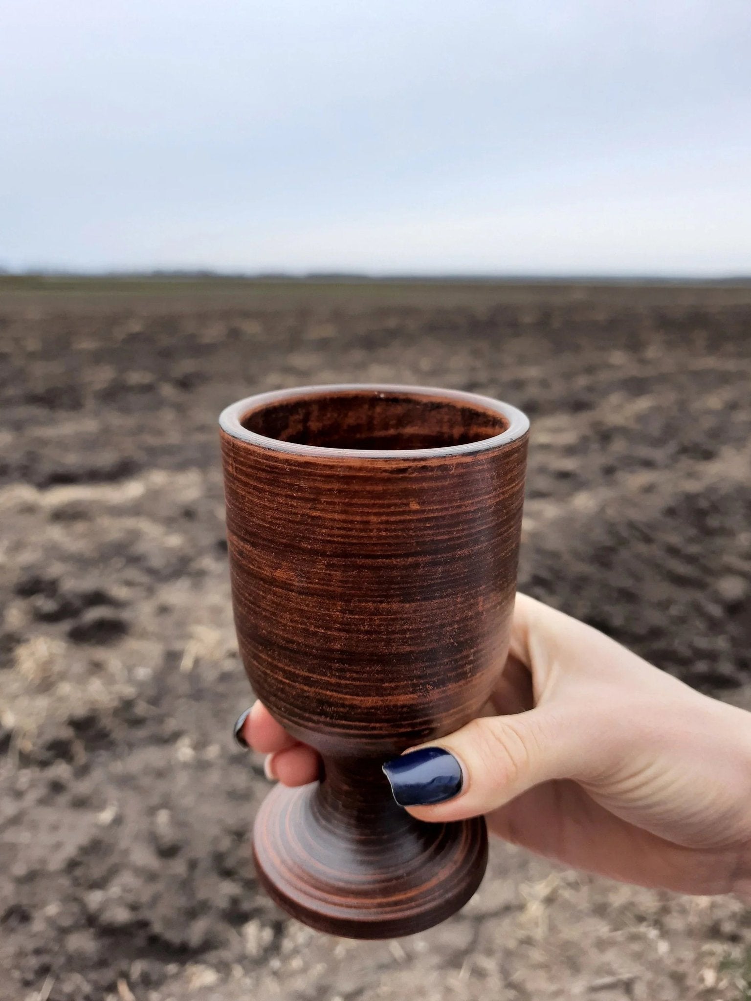 Hand holding a handmade clay wine glass with a rustic brown finish outdoors